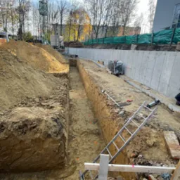 Wykop pod fundamenty z widocznymi znacznikami geodezyjnymi, drabina oparta o skarpę, w tle betonowy mur oporowy i drzewa.
