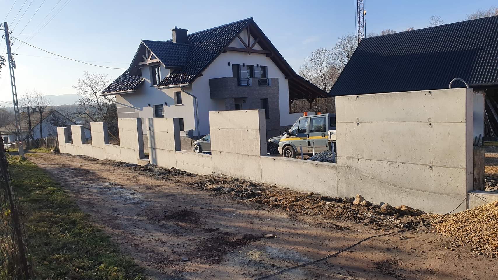 Wznoszenie betonowego muru oporowego na nierównym terenie, z widocznym samochodem dostawczym i fragmentem domu w tle.