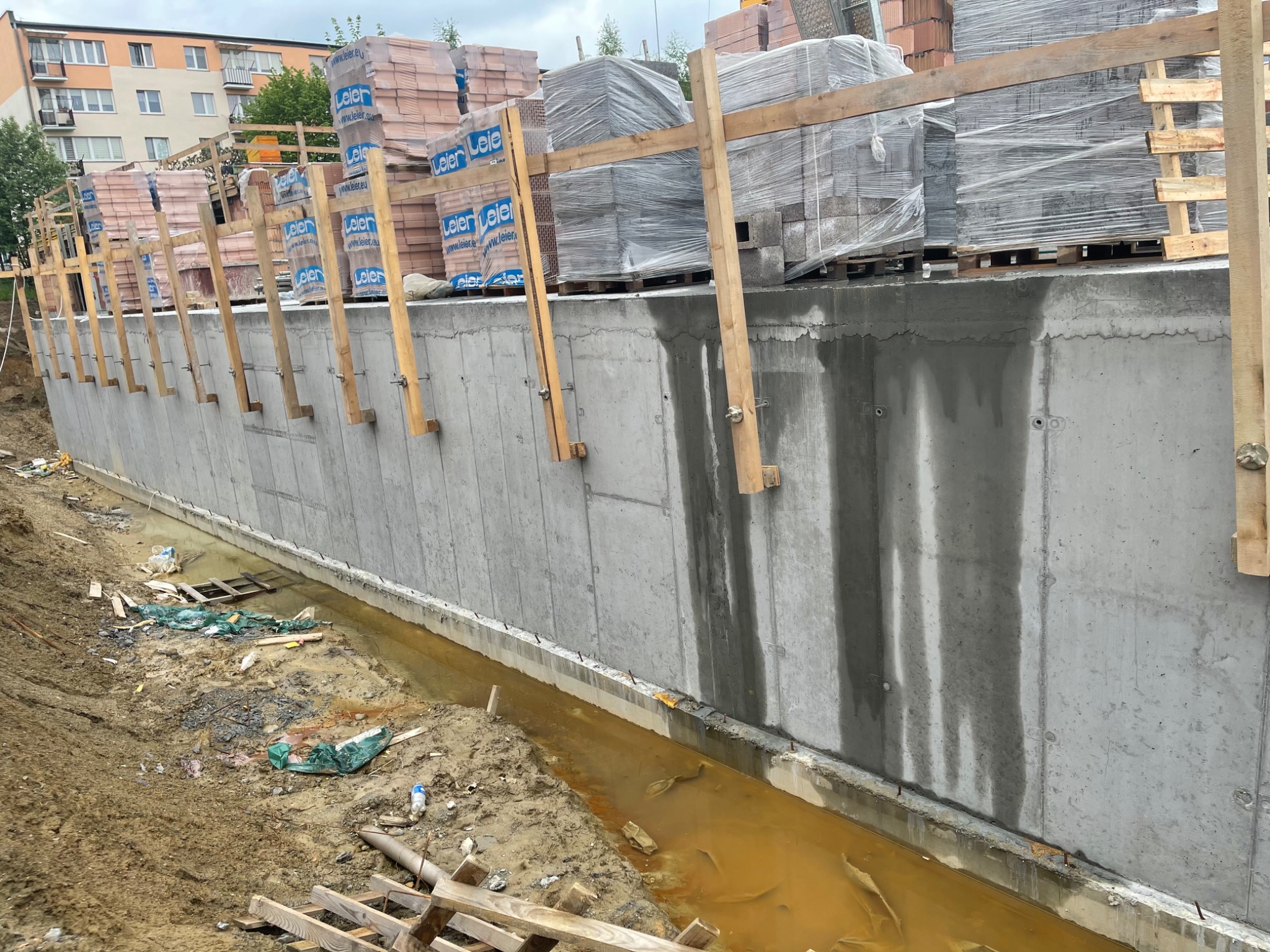 Wzniesiony betonowy fundament z drewnianym szalunkiem i zapasem materiałów budowlanych, w wykopie z wodą i resztkami budowlanymi.