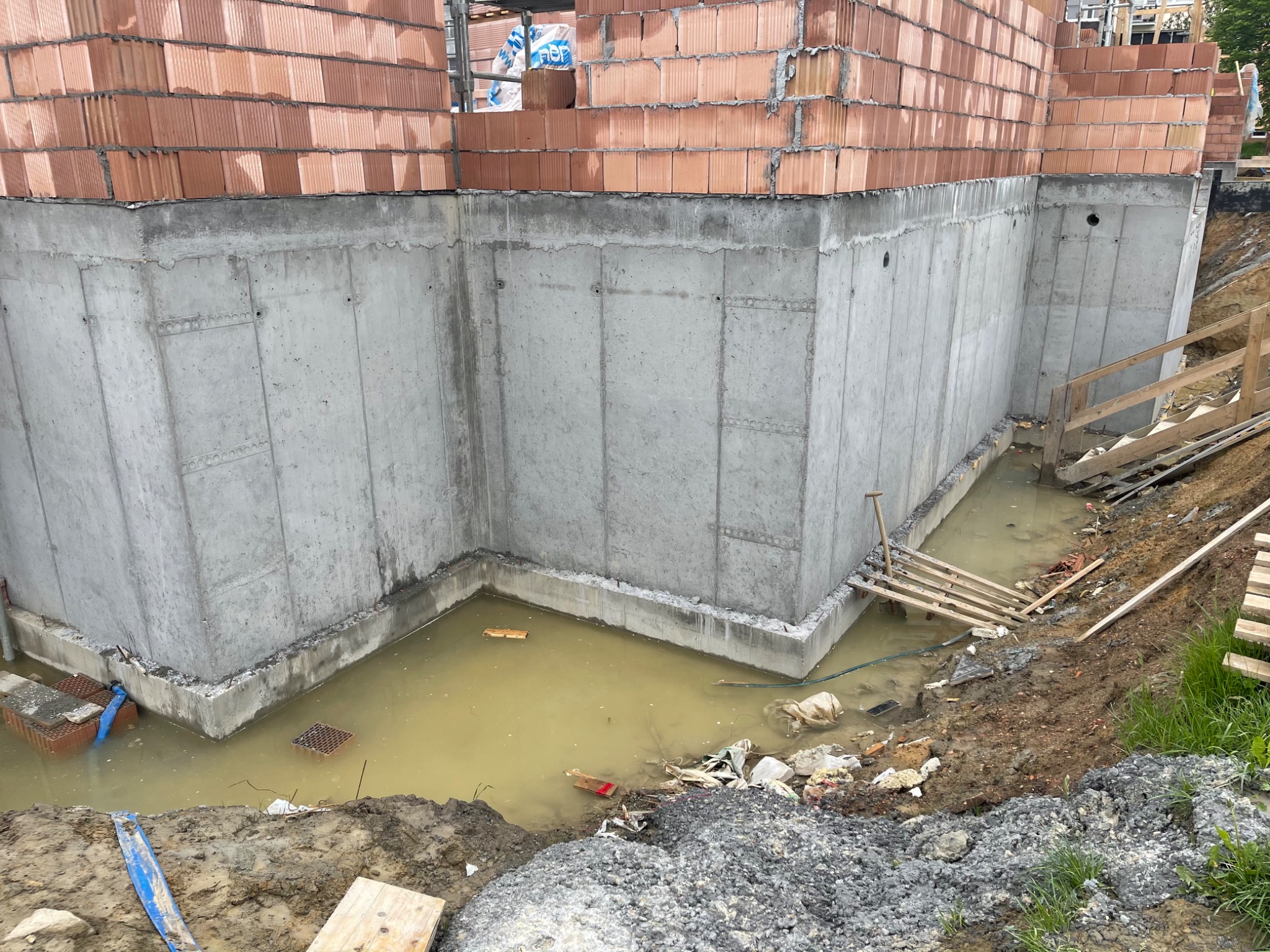 Surowy betonowy fundament budynku w trakcie budowy, częściowo zalany wodą, z widocznymi elementami konstrukcyjnymi i gruzem budowlanym w wykopie.