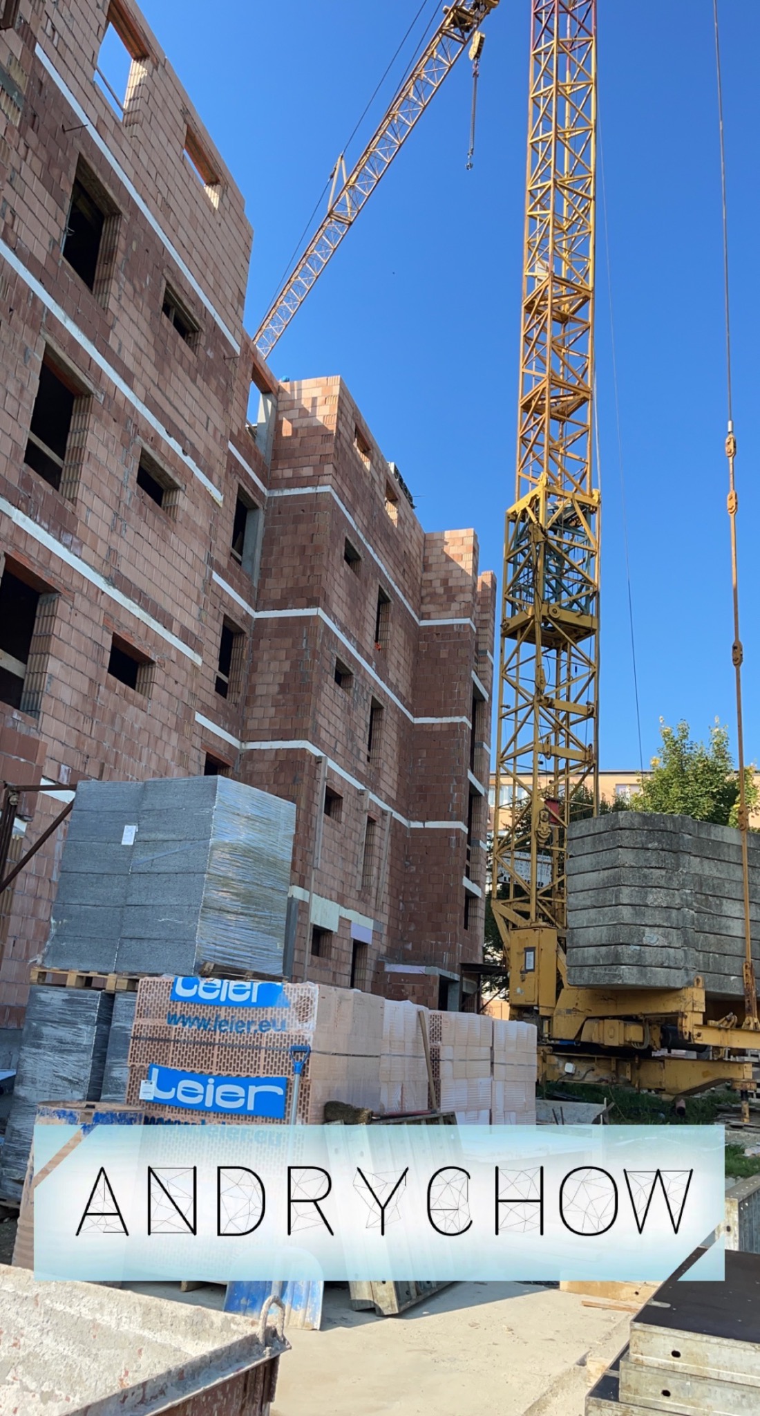 Wznoszenie wielopiętrowego budynku z cegły ceramicznej, widoczny żółty dźwig budowlany i palety z materiałami budowlanymi marki Leier.