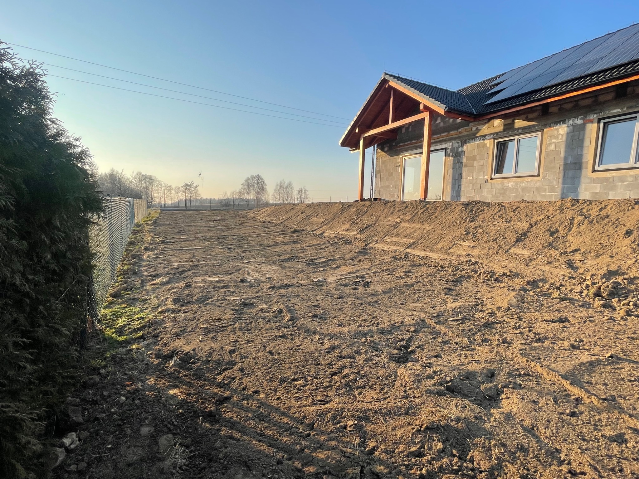 Surowy stan domu z pustaków z drewnianą konstrukcją dachu i panelami słonecznymi, widok na przygotowany teren z ziemią, ogrodzenie z siatki i drzewa w tle.