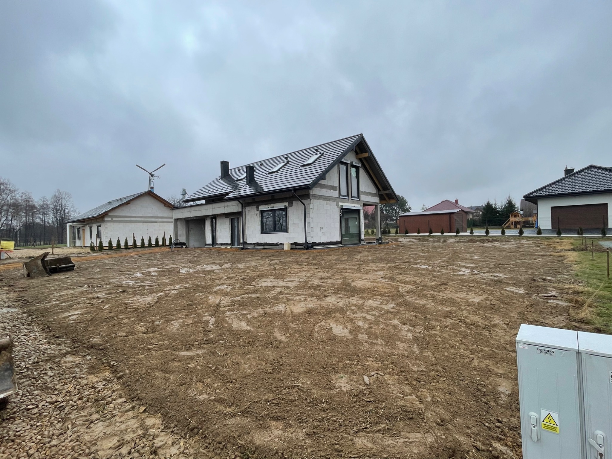 Surowy stan domu murowanego z czarną dachówką i oknami, widoczne elementy szalunku, teren wokół wyrównany, dzień pochmurny.