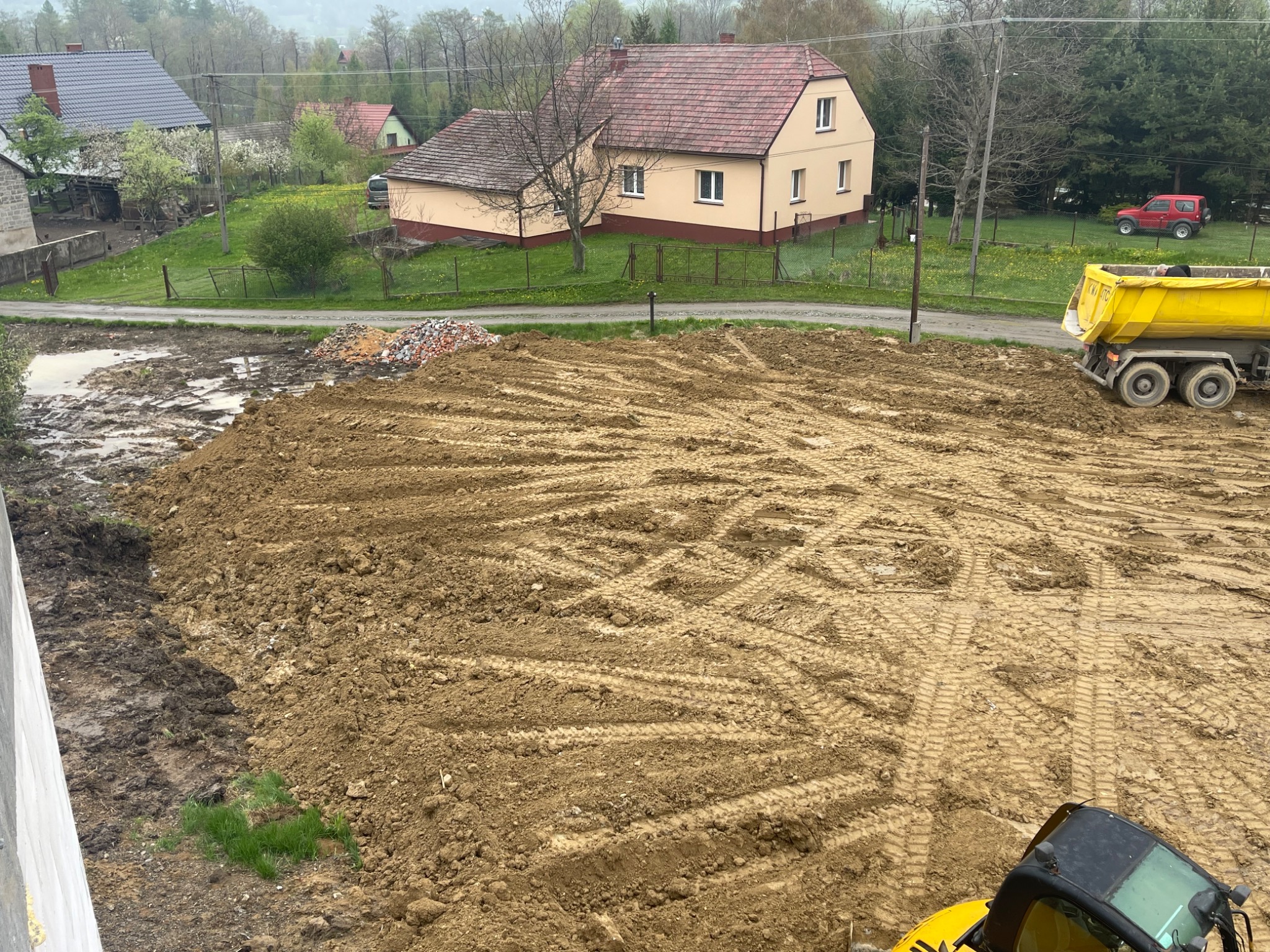 Przygotowany teren pod budowę z widocznym żółtym wywrotką i fragmentem koparki, ślady opon na ubitej ziemi, w tle zabudowa wiejska i drzewa.