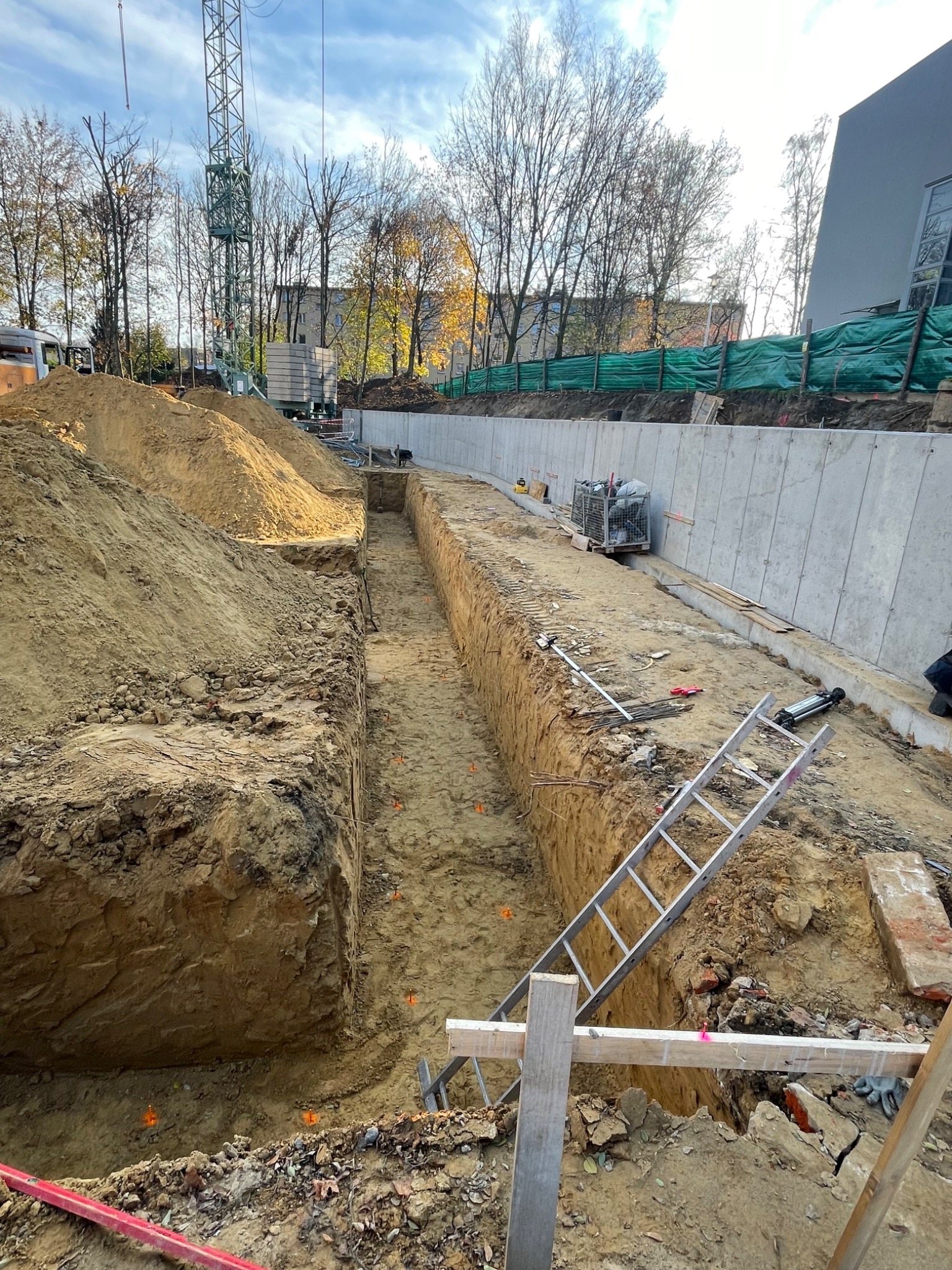 Wykop pod fundamenty z widocznymi znacznikami geodezyjnymi, drabina oparta o skarpę, w tle betonowy mur oporowy i drzewa.