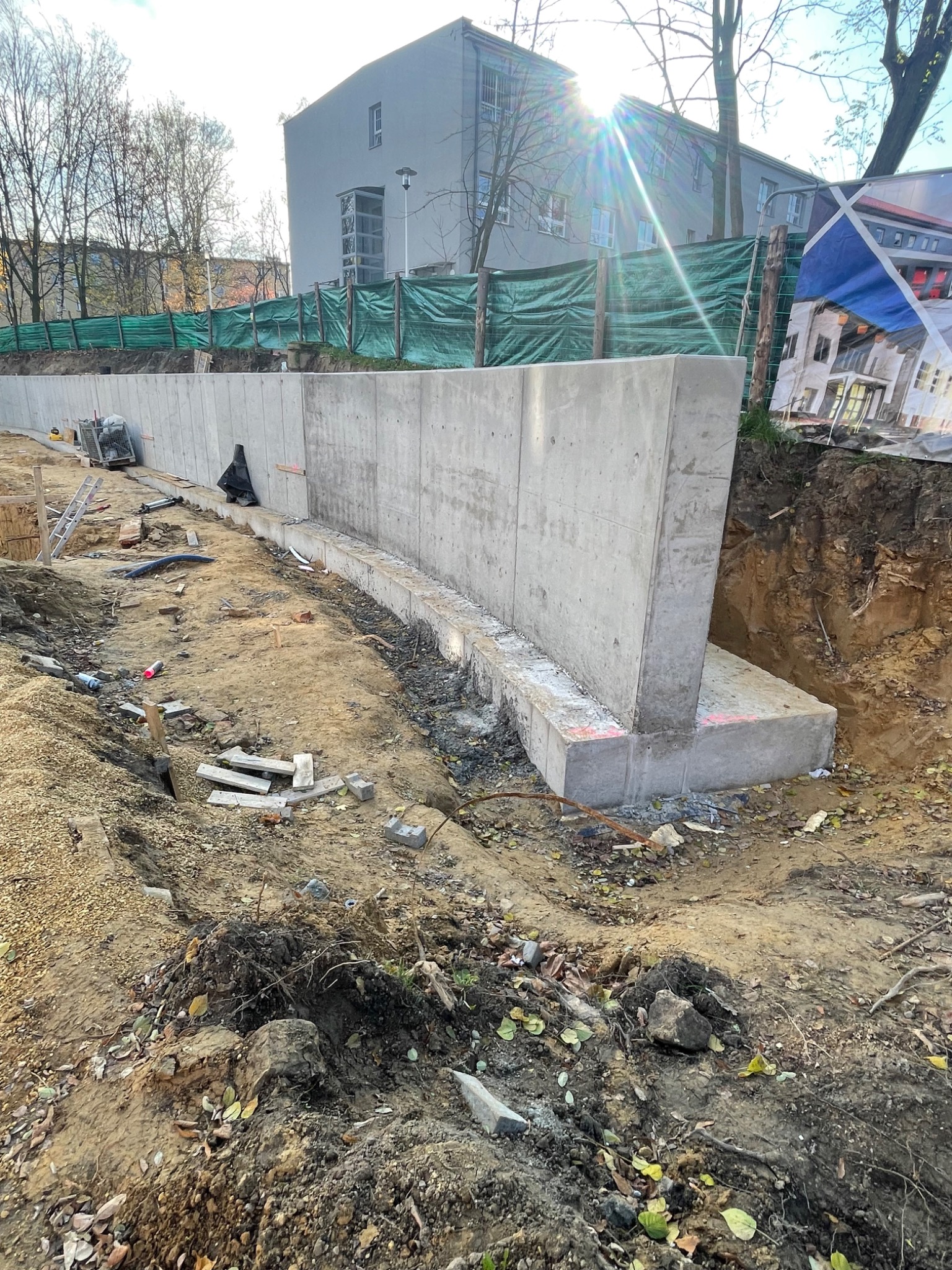 Betonowa ściana oporowa w trakcie budowy, widoczne fundamenty, ziemia i narzędzia budowlane, w tle budynek z zieloną siatką ochronną i reklama budynku.