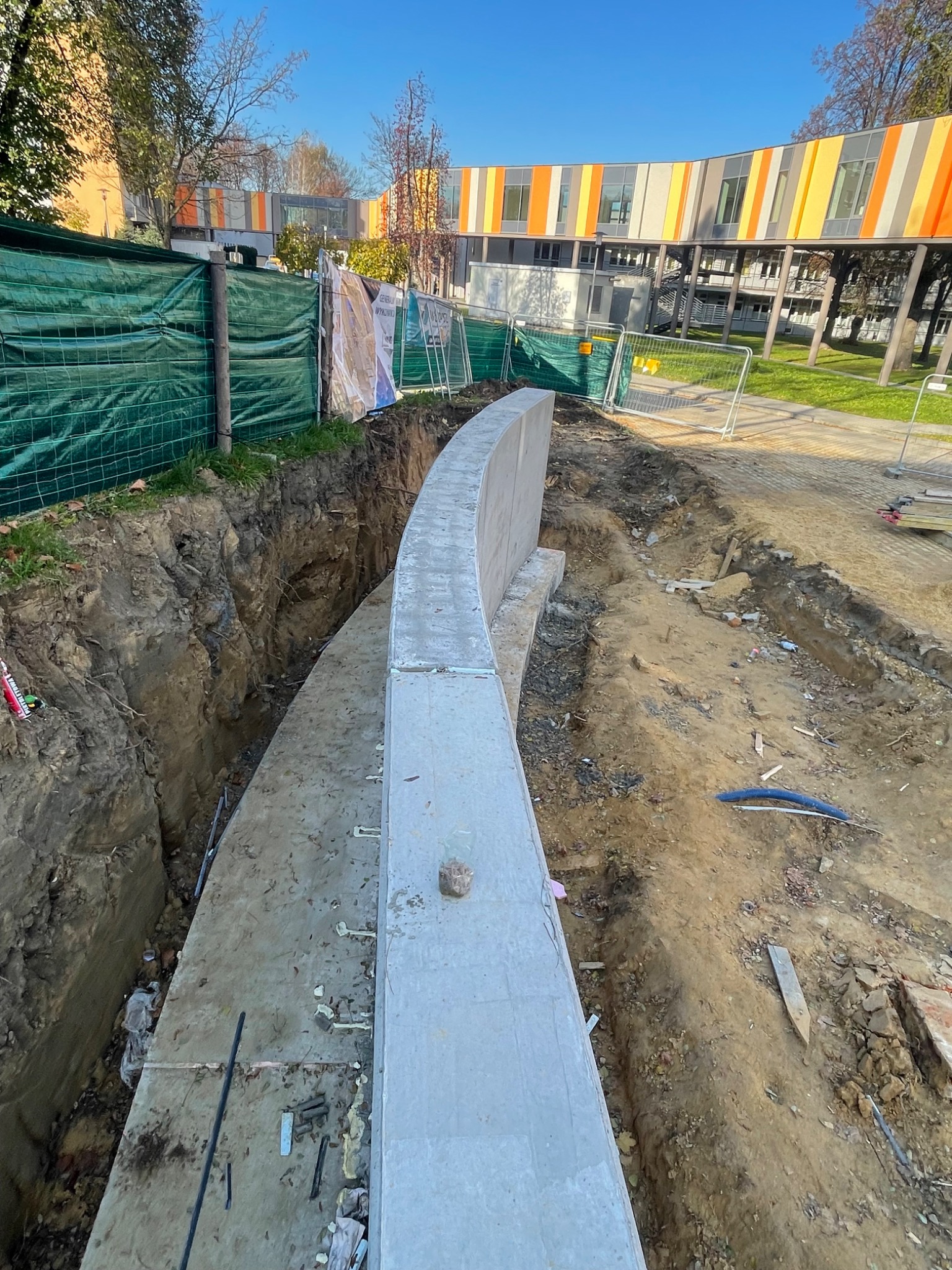 Wykop fundamentowy pod zakrzywioną ścianę oporową na budowie, widoczne zbrojenie, ziemia i w tle budynek z kolorowymi panelami elewacyjnymi.