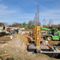 Montaż szalunków i zbrojenia fundamentów pod budynek na placu budowy; widoczny dźwig teleskopowy i ciężarówka z pracownikami.