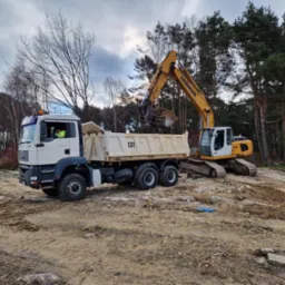 Żółta koparka Liebherr 914 ładuje ziemię do białej ciężarówki MAN TGA 41.480 na placu budowy z lasem w tle.