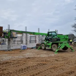 Budowa domu z betonu komórkowego: zielony podnośnik teleskopowy ROTO 33.16 z pracownikami na platformie, widoczne zbrojenia i palety z bloczkami, teren budowy w błocie.