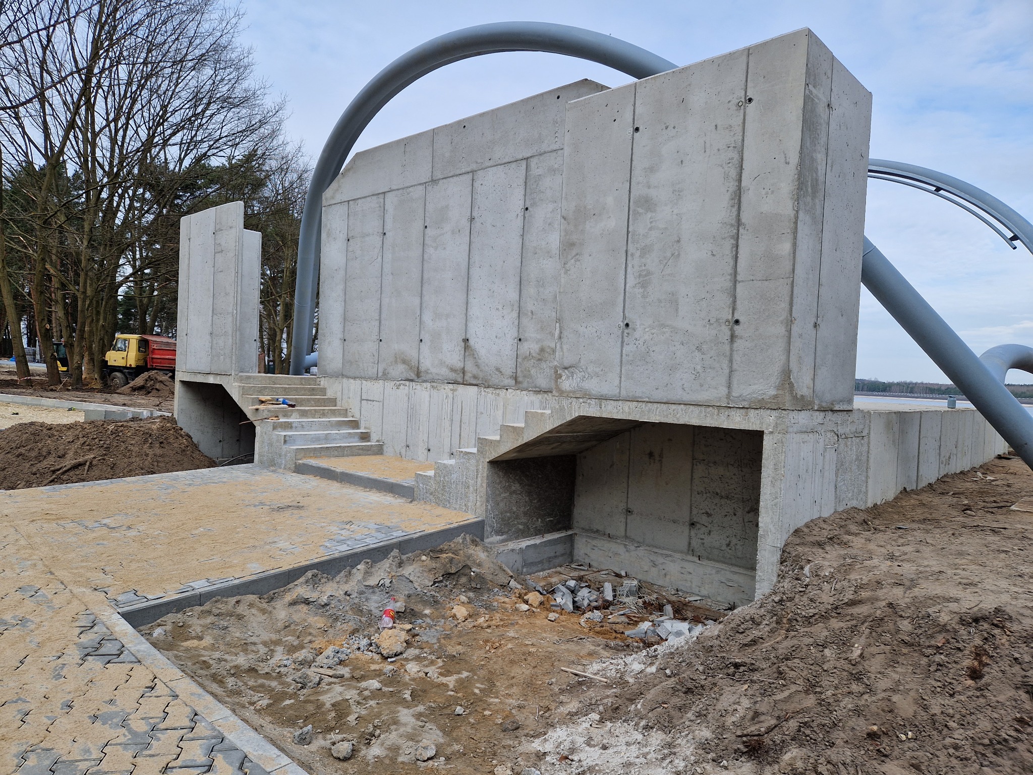 Surowa betonowa konstrukcja z schodami w trakcie budowy, widoczne ślady prac ziemnych i przygotowanie terenu, w tle żółta ciężarówka i drzewa.