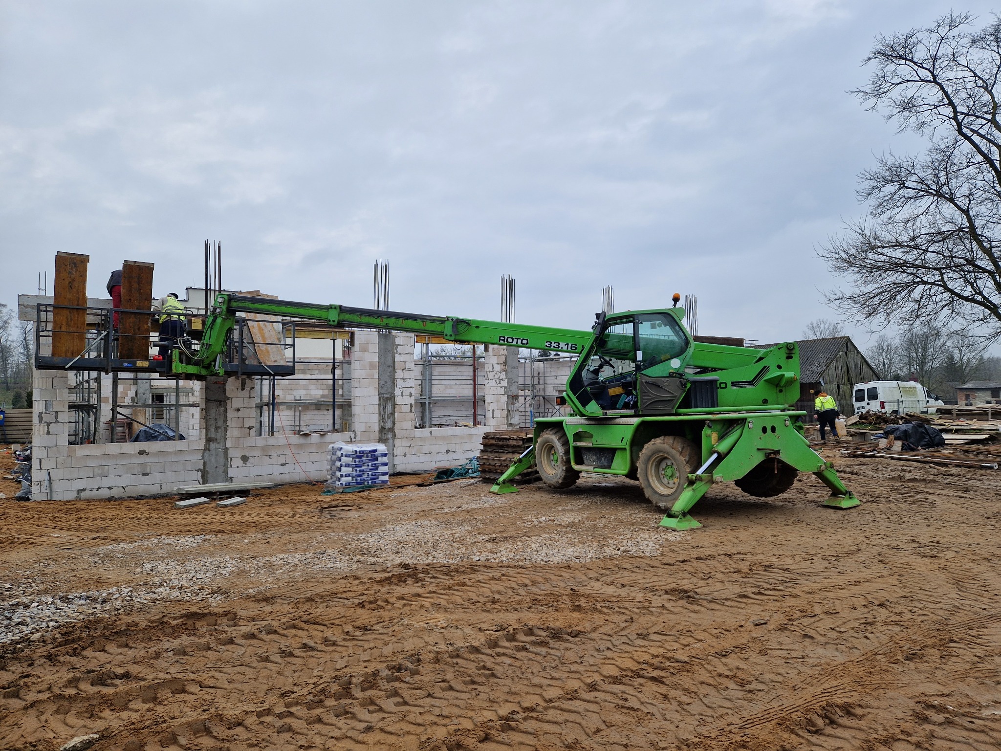 Budowa domu z betonu komórkowego: zielony podnośnik teleskopowy ROTO 33.16 z pracownikami na platformie, widoczne zbrojenia i palety z bloczkami, teren budowy w błocie.