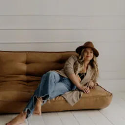 Kobieta w brązowym kapeluszu i beżowym płaszczu relaksuje się na sk&oacute;rzanej sofie w jasnym, minimalistycznym wnętrzu z białymi panelami ściennymi i podłogą.