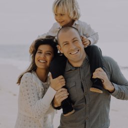 Alicja Kupczyk Fotografia - Uśmiechnięta rodzina: mama w ażurowej sukience, tata z synkiem na barana, na tle plaży i morza.