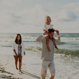 Alicja Kupczyk Fotografia - Uśmiechnięta rodzina spaceruje brzegiem morza w słoneczny dzień: tata z córeczką na barana, mama idzie obok.