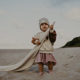 Alicja Kupczyk Fotografia - Dziewczynka w beżowej czapce i różowej sukience stoi na piaszczystej plaży, trzymając w rękach kremowy, teksturowany kocyk.