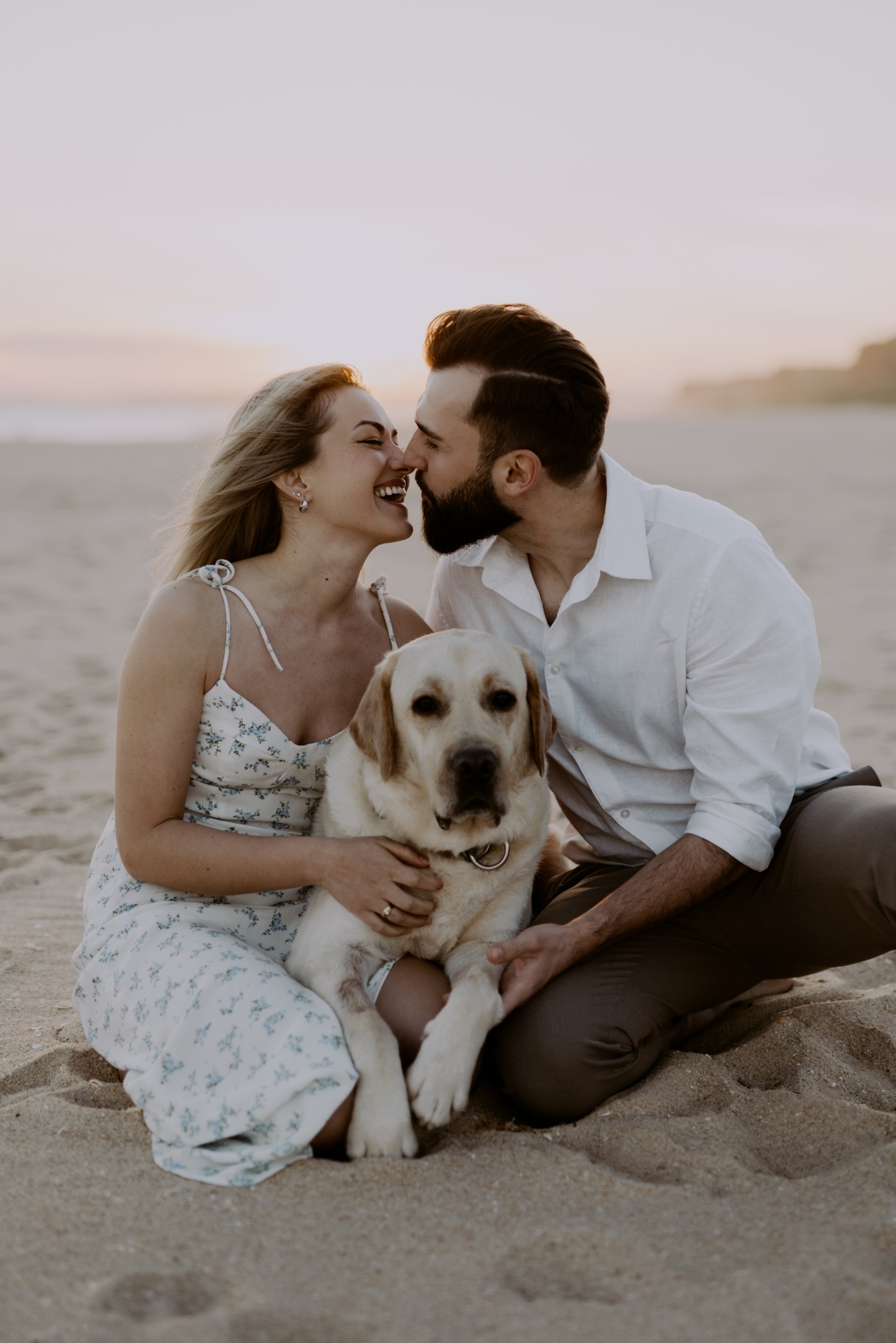 Para zakochanych na plaży, dotykają się nosami, siedzą z psem labrador retriever o jasnej sierści, zachód słońca w tle.