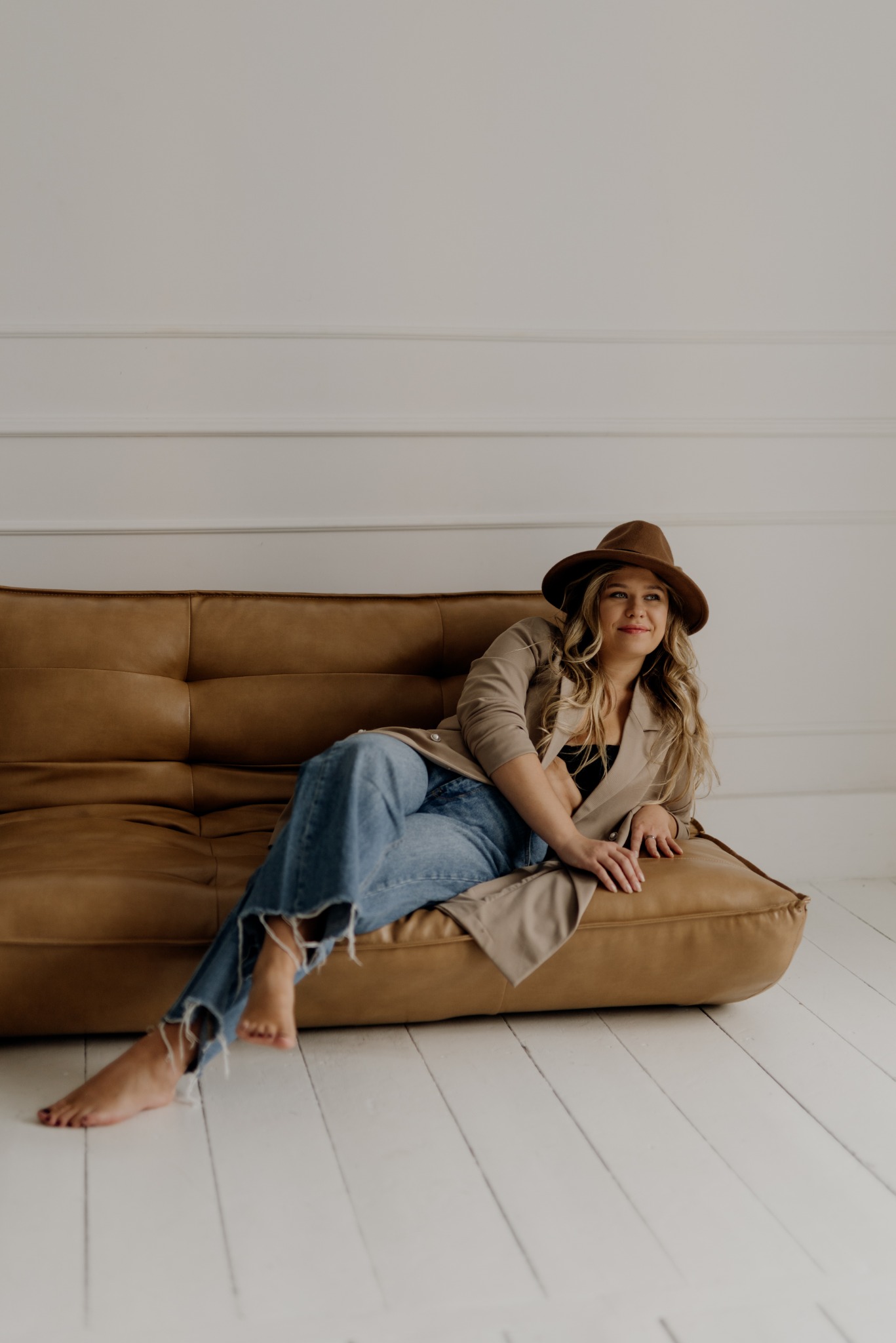 Kobieta w brązowym kapeluszu i beżowym płaszczu relaksuje się na skórzanej sofie w jasnym, minimalistycznym wnętrzu z białymi panelami ściennymi i podłogą.
