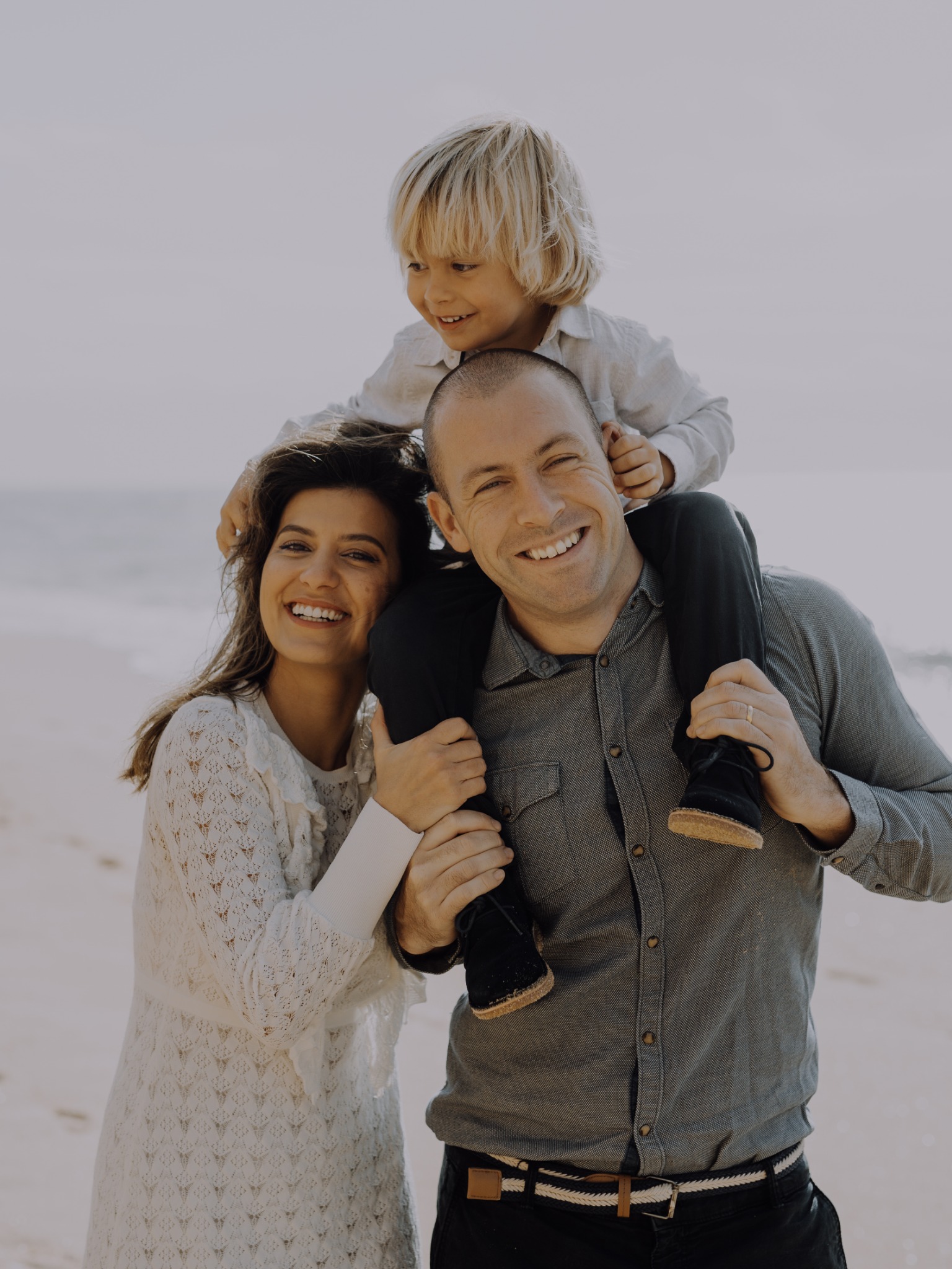 Uśmiechnięta rodzina: mama w ażurowej sukience, tata z synkiem na barana, na tle plaży i morza.