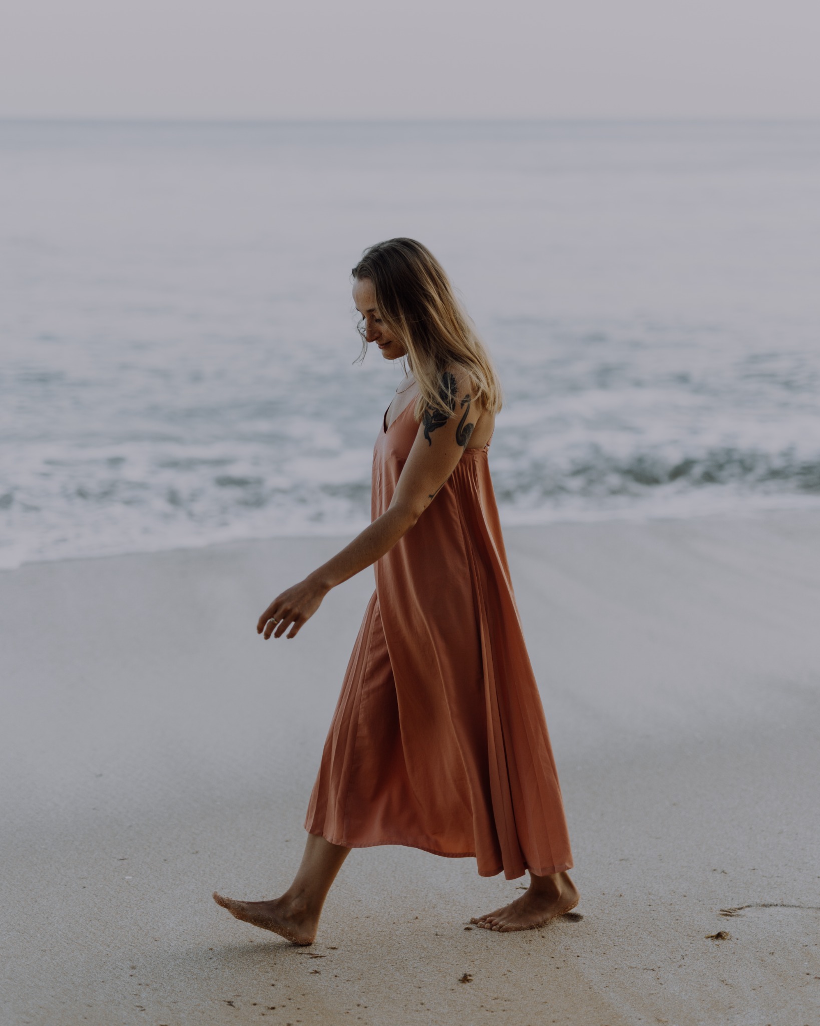 Kobieta w łososiowej sukience spaceruje boso po plaży w miękkim, rozproszonym świetle poranka, z tatuażem na ramieniu i delikatnymi falami oceanu w tle.