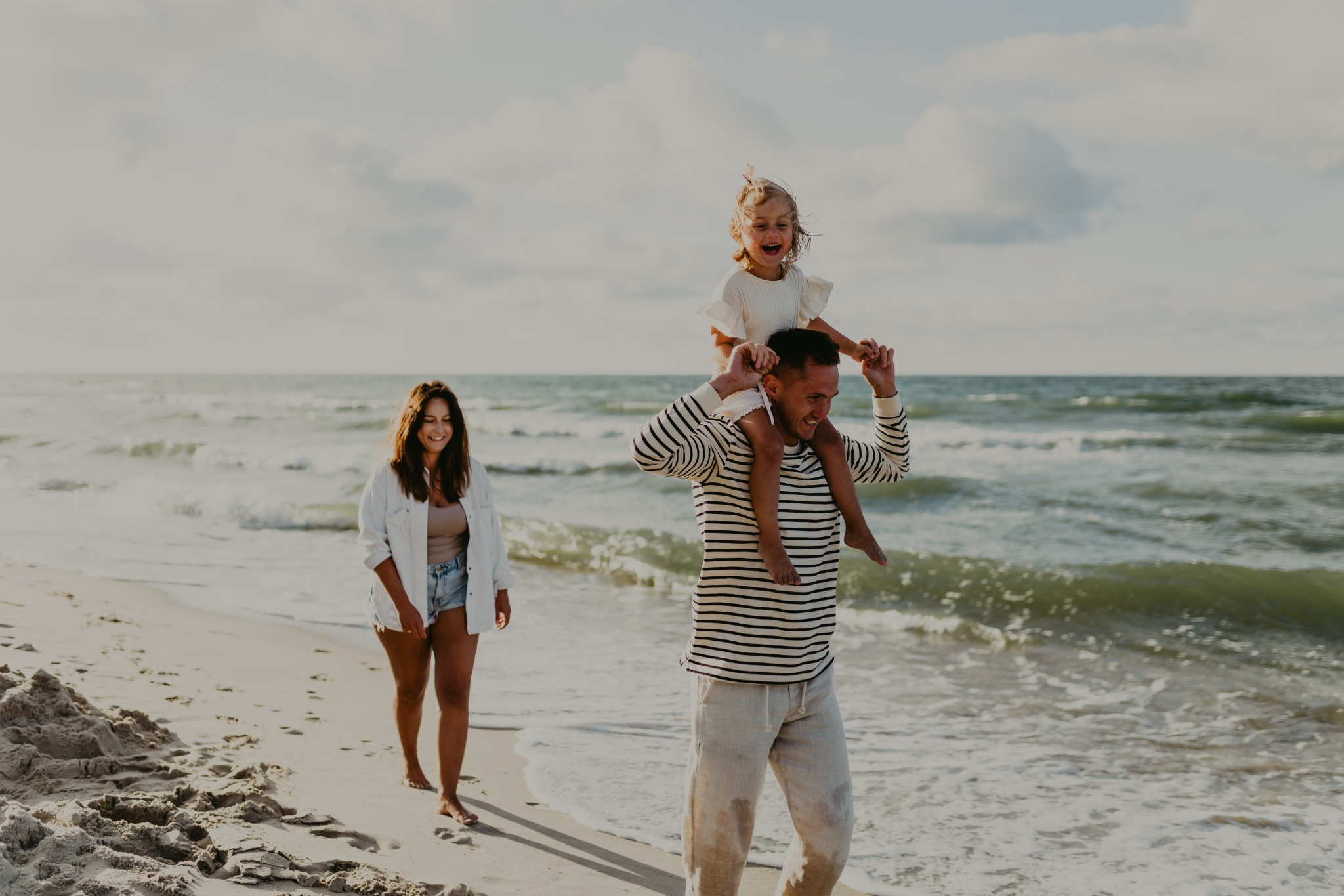 Uśmiechnięta rodzina spaceruje brzegiem morza w słoneczny dzień: tata z córeczką na barana, mama idzie obok.