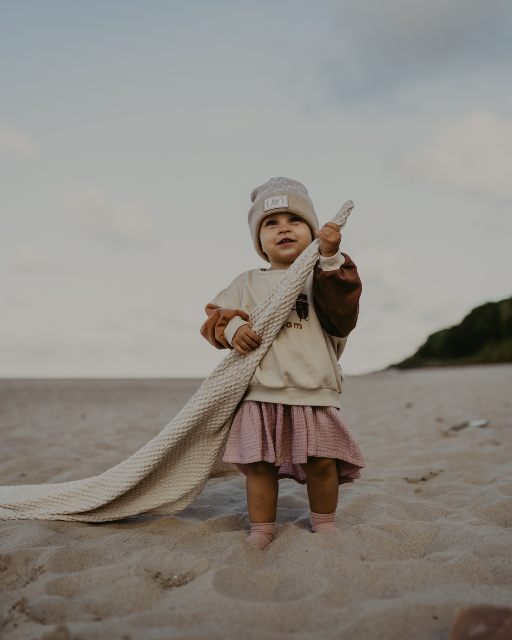 Dziewczynka w beżowej czapce i różowej sukience stoi na piaszczystej plaży, trzymając w rękach kremowy, teksturowany kocyk.