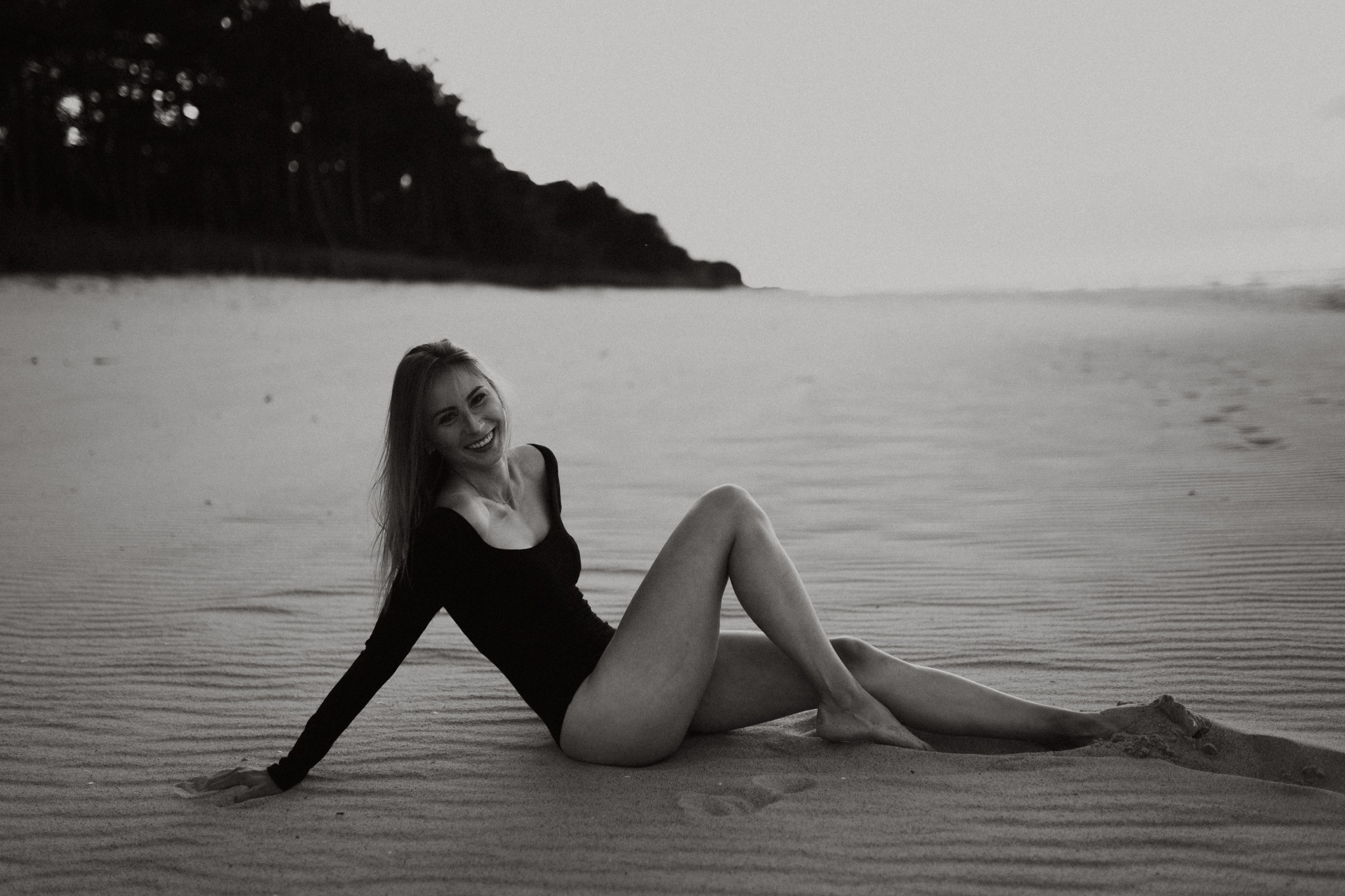 Uśmiechnięta kobieta w czarnym stroju kąpielowym leży na piaszczystej plaży w monochromatycznej tonacji, z widocznymi śladami stóp na piasku i zalesionym wzgórzem w tle.