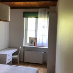 Pokój z drewnianym sufitem, białymi ścianami, biurkiem i łóżkiem, oświetlony światłem dziennym wpadającym przez okno z zieloną roletą.