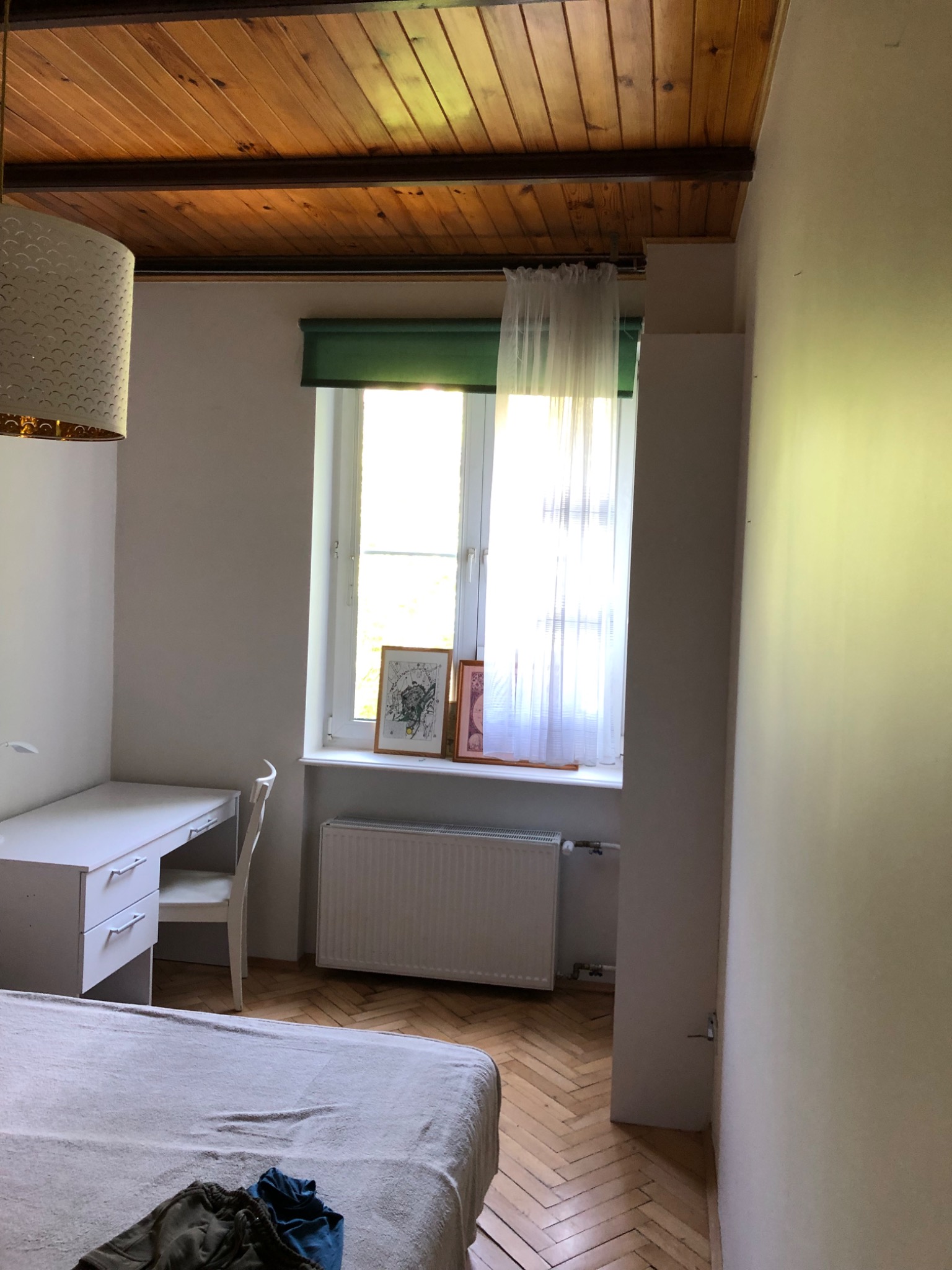 Pokój z drewnianym sufitem, białymi ścianami, biurkiem i łóżkiem, oświetlony światłem dziennym wpadającym przez okno z zieloną roletą.