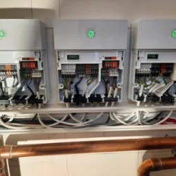 Trzy moduły rozszerzające instalację grzewczą MM-100, każdy z zieloną diodą sygnalizacyjną, widoczne liczne przewody elektryczne i rury miedziane, zamontowane na białej ścianie.