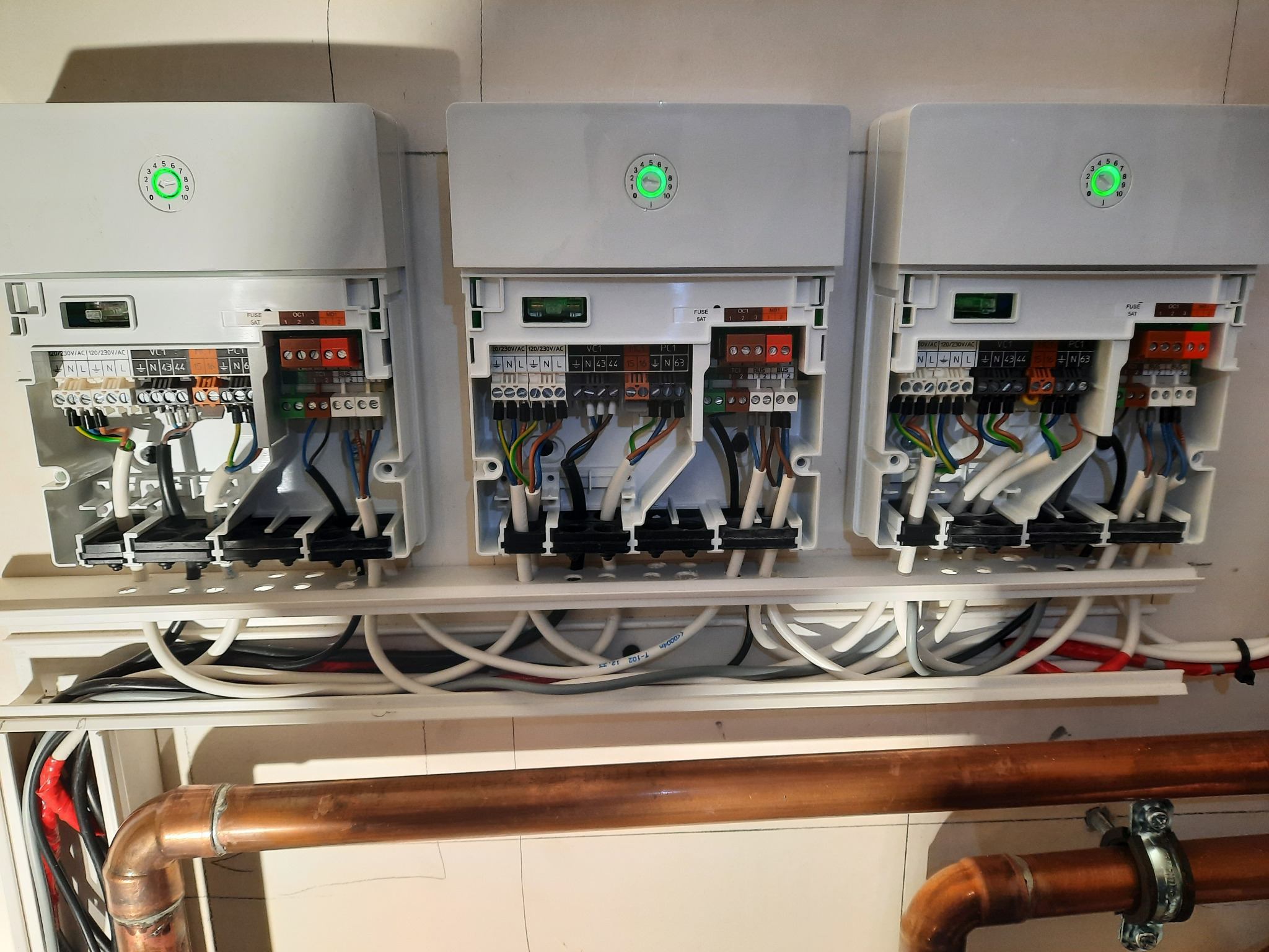 Trzy moduły rozszerzające instalację grzewczą MM-100, każdy z zieloną diodą sygnalizacyjną, widoczne liczne przewody elektryczne i rury miedziane, zamontowane na białej ścianie.