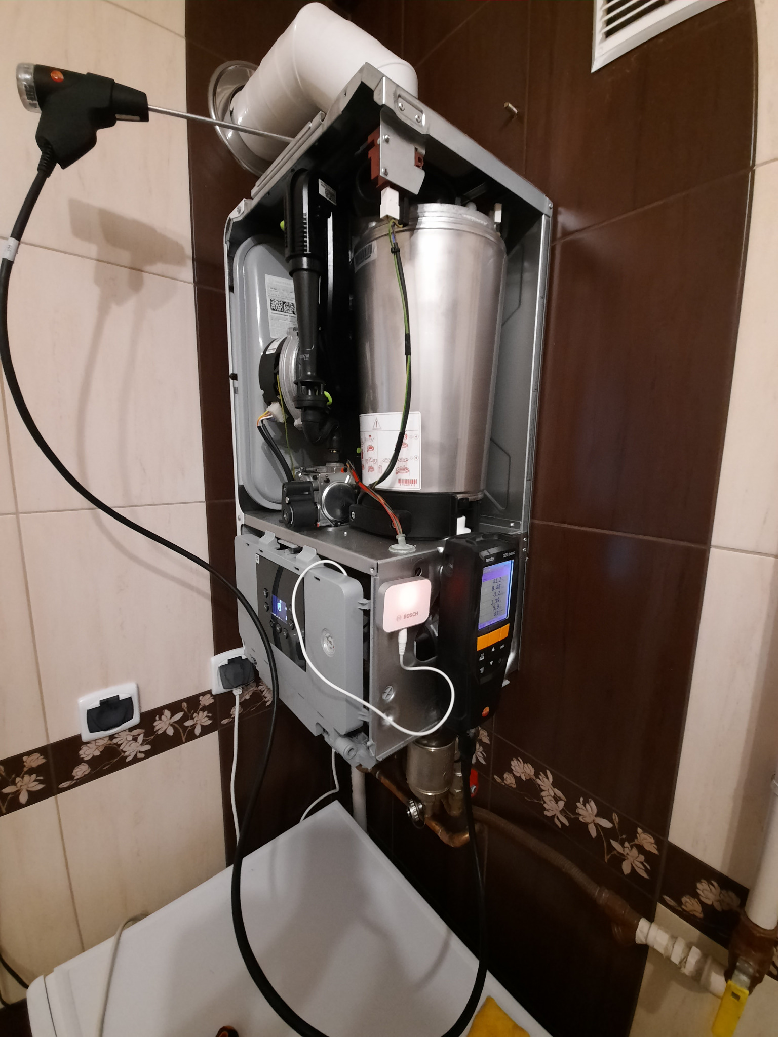 Otwarty piec gazowy z podłączonym analizatorem spalin marki Testo, widoczne parametry na wyświetlaczu, tło z płytek ceramicznych.