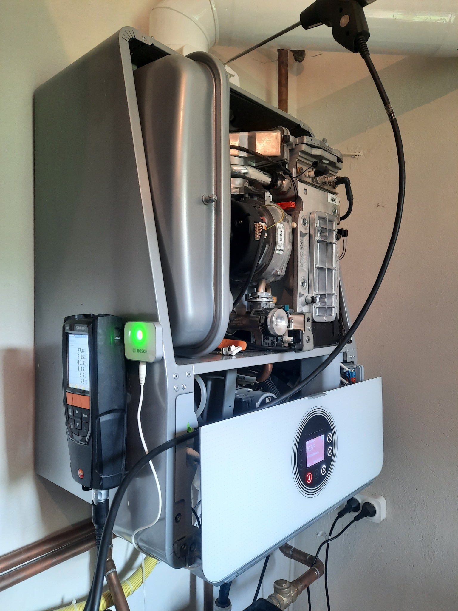 Otwarty piec gazowy Bosch Condens GC9000iW z podłączonym analizatorem spalin i czujnikiem Bosch, widoczne rury miedziane i żółty przewód gazowy.