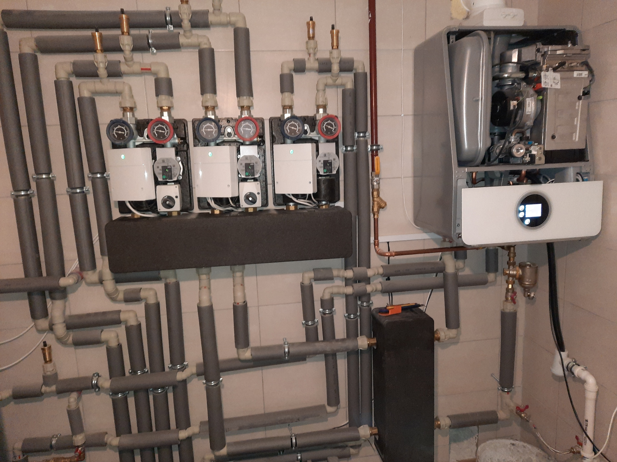 Zainstalowany piec gazowy Bosch Condens GC9000iW z rozbudowaną instalacją hydrauliczną, widoczne liczne rury z izolacją termiczną, manometry i zawory, całość na tle kafelkowanej ściany.