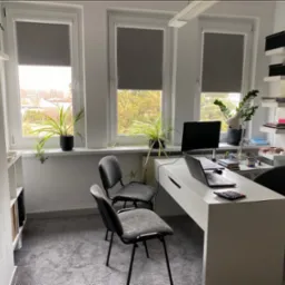 Jasne biuro z trzema oknami z roletami, biurkiem z laptopem i kalkulatorem, dwoma krzesłami dla gości i obrotowym krzesłem biurowym, roślinami doniczkowymi na parapetach i ścianą z zegarem.