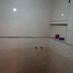 Ściana łazienki wyłożona beżowymi płytkami ceramicznymi z dekoracyjnym pasem, widoczne przyłącza wodne i odpływ kanalizacyjny w trakcie prac wykończeniowych.