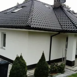 Nowoczesny dom jednorodzinny z ciemną dachówką ceramiczną, białymi ścianami z ciemnym cokołem, oknami dachowymi i zadbanym trawnikiem, widoczny z boku.