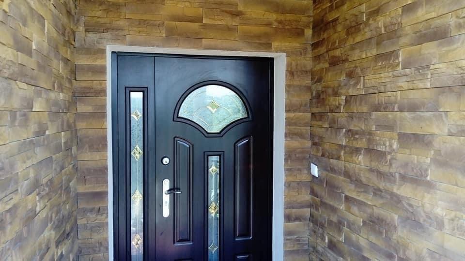 Ciemne drzwi wejściowe z ozdobnymi szybami, obramowane imitacją kamienia w odcieniach brązu i beżu.