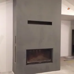 Nowoczesny, szary kominek w trakcie montażu, widoczne narzędzia i akcesoria na surowej podłodze, surowe drewniane belki stropowe.