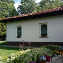 Biały parterowy dom z brązowym cokołem i czerwoną dachówką, okna z firankami i kwiatami, rabata z kwiatami i krzewami, skrzynka z żółtymi bratkami, konewka.