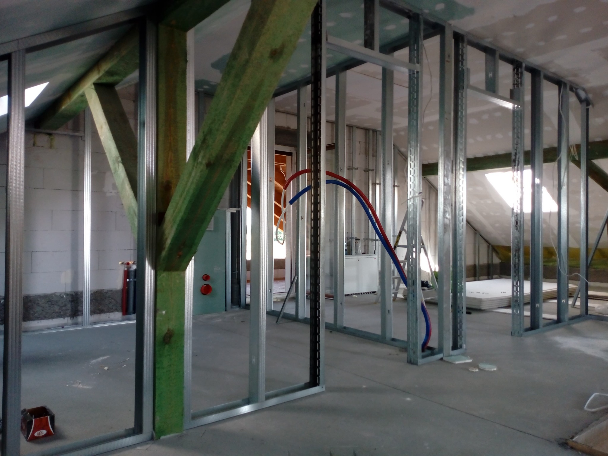 Szkielet ścian działowych z profili metalowych w surowym stanie poddasza, widoczne instalacje wodne, drewniany słup nośny, okna dachowe i przygotowane płyty gipsowo-kartonowe.