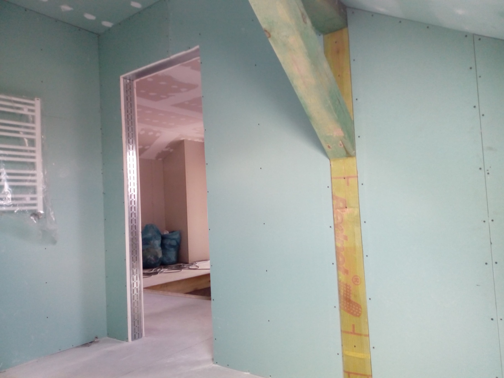 Montaż płyty karton gips na ścianie z widoczną konstrukcją drewnianą i ociepleniem, widok na sąsiednie pomieszczenie przez metalową futrynę drzwiową, grzejnik pod folią w tle.