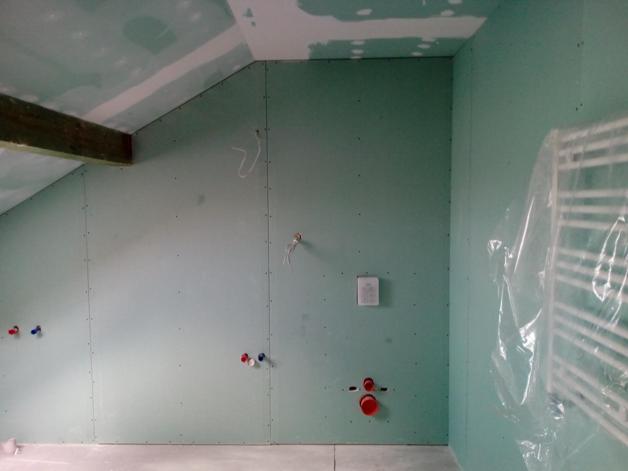 Pokój na poddaszu w trakcie wykańczania: ściany i sufit pokryte płytami gipsowo-kartonowymi, widoczne instalacje wodne, drewniana belka stropowa i grzejnik owinięty folią ochronną.