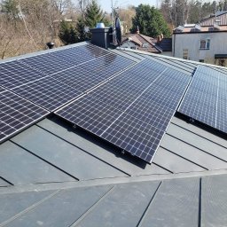 Świat Miodu Sp. z o.o. - Panele słoneczne zamontowane na szarym dachu z blachy trapezowej, widoczny komin z anteną satelitarną, w tle budynki i drzewa.