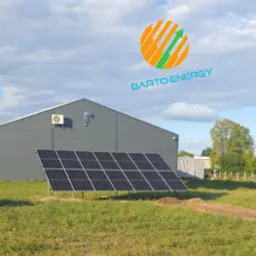 Instalacja paneli słonecznych na trawiastym terenie przed szarym budynkiem magazynowym, widoczne logo firmy na tle pochmurnego nieba.