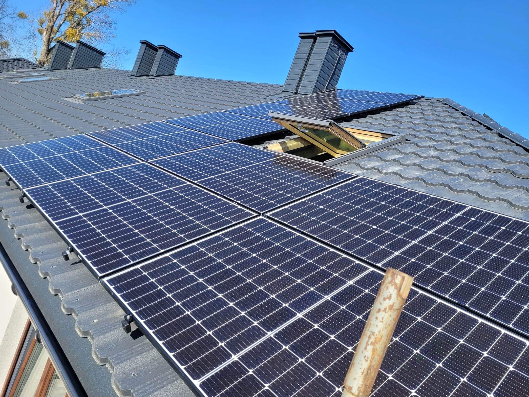 Panele słoneczne na szarym dachu z dachówką, widoczne okno dachowe i kominy, fragment drabiny w prawym dolnym rogu.