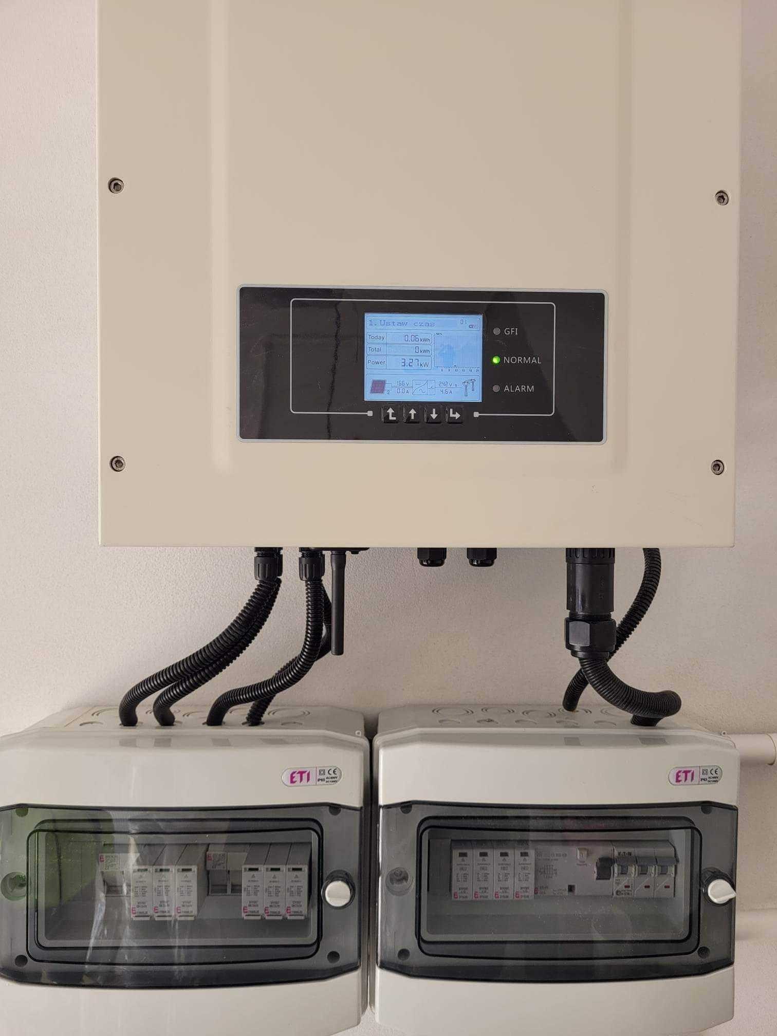 Biała skrzynka inwertera fotowoltaicznego z wyświetlaczem pokazującym dane produkcji energii, pod nią dwie szare skrzynki zabezpieczeń ETI, kable w czarnych peszlach.