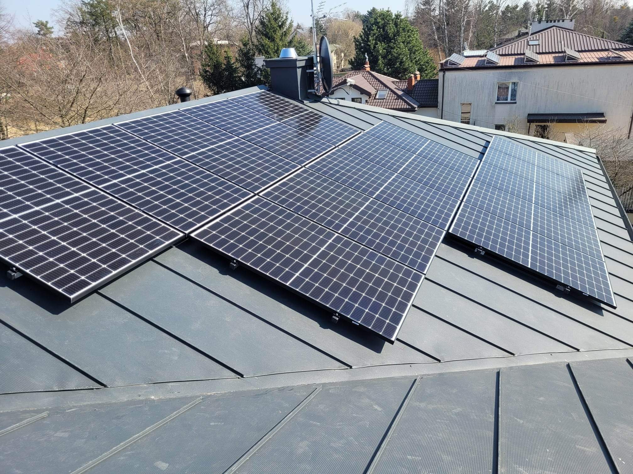 Panele słoneczne zamontowane na szarym dachu z blachy trapezowej, widoczny komin z anteną satelitarną, w tle budynki i drzewa.