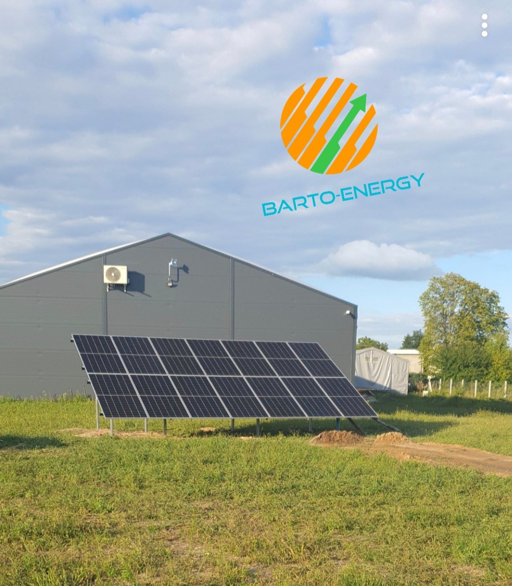 Instalacja paneli słonecznych na trawiastym terenie przed szarym budynkiem magazynowym, widoczne logo firmy na tle pochmurnego nieba.