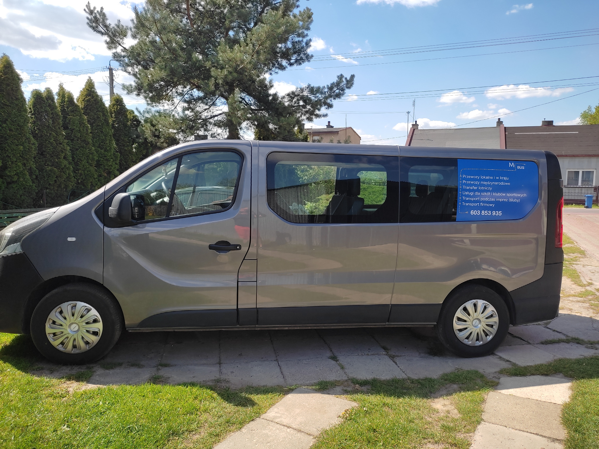 Szary bus z niebieską naklejką reklamową na szybie, oferujący przewozy lokalne, międzynarodowe, transfer lotniczy, usługi dla szkół i transport na imprezy, zaparkowany na trawie przed domem.